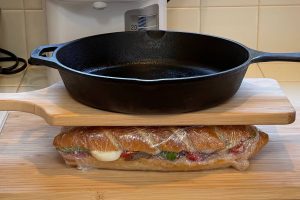 sandwich wrapped in plastic wrap being pressed by heavy cast iron pan