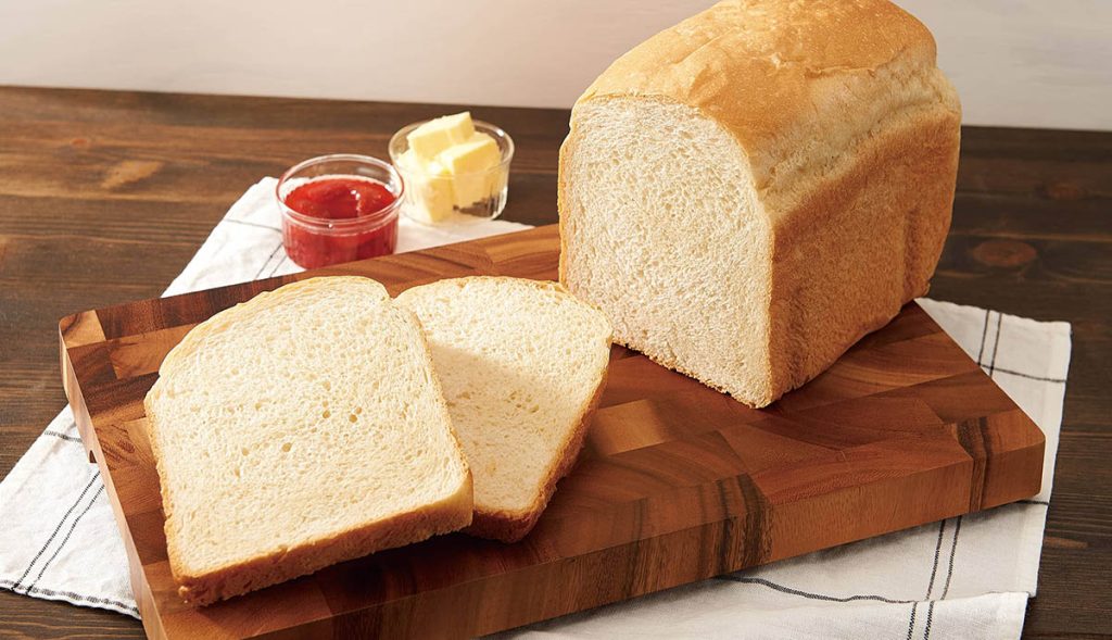 A loaf of white bread with a couple of slices cut on a wooden cutting board. Butter and jam are on the side.