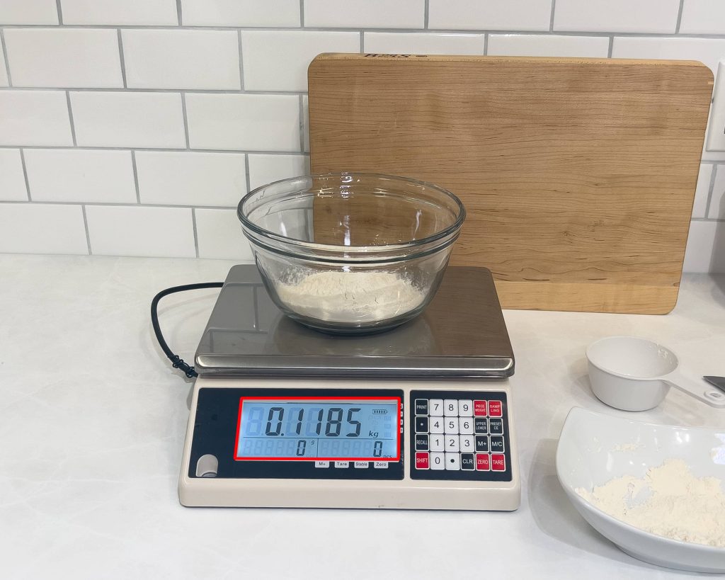 A cup of fluffed flour is in a glass bowl on a kitchen scale and it reads 118 grams