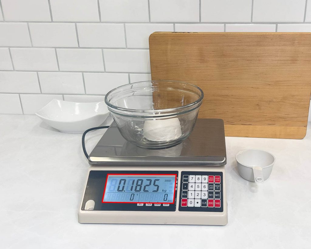 A cup of packed flour in a glass bowl on a kitchen scale which is measuring a weight of 182 grams