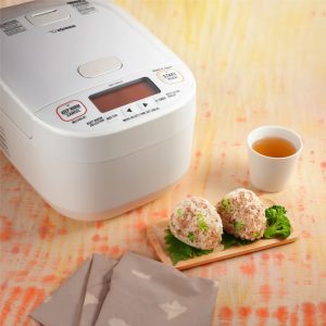 2 onigiri next to a white ricecooker and hot tea