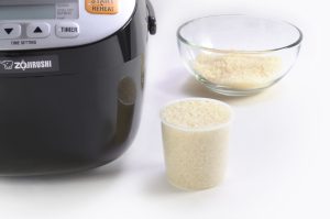 a cup of uncooked rice and rice cooker in a white background